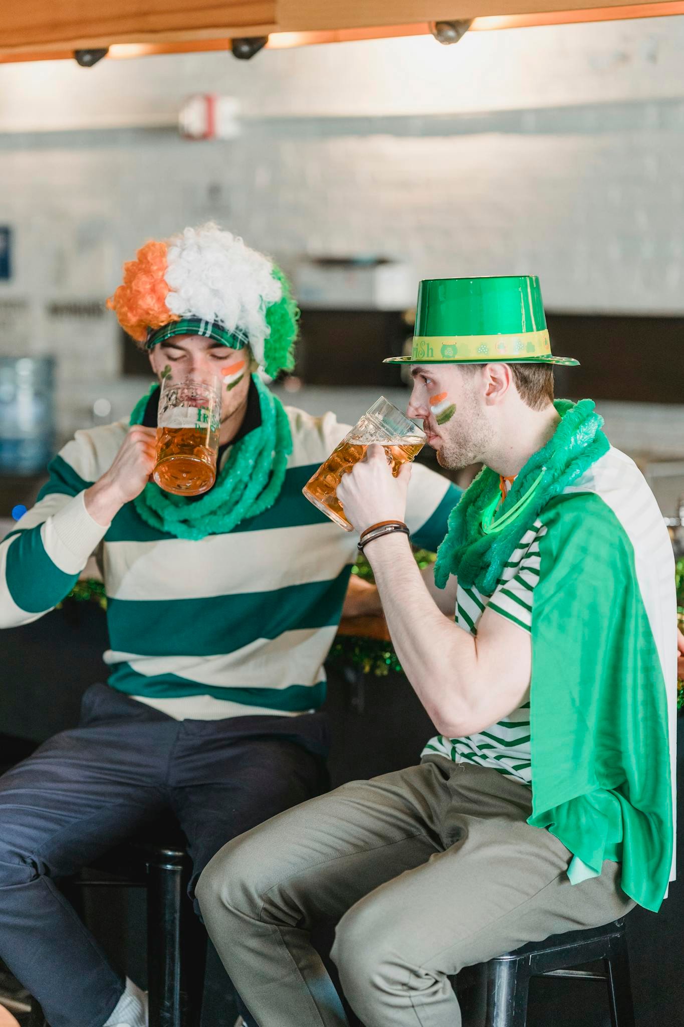 Male friends in traditional clothes and hats drinking beer while sitting on chairs at counter in cafe during Saint Patricks day